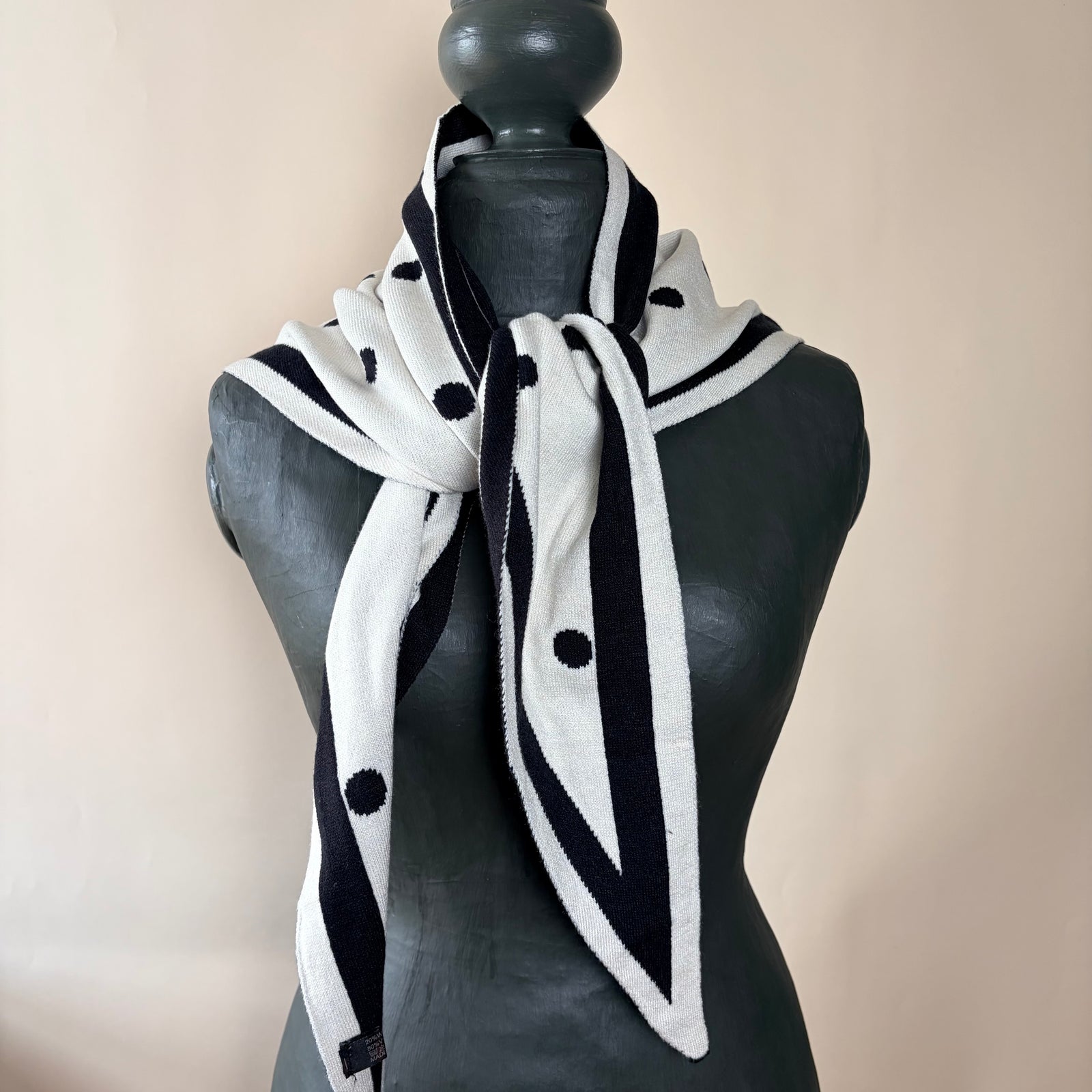 A black and white knit scarf with white polka dots and striped trim, styled on a dark gray mannequin torso against a light beige background.