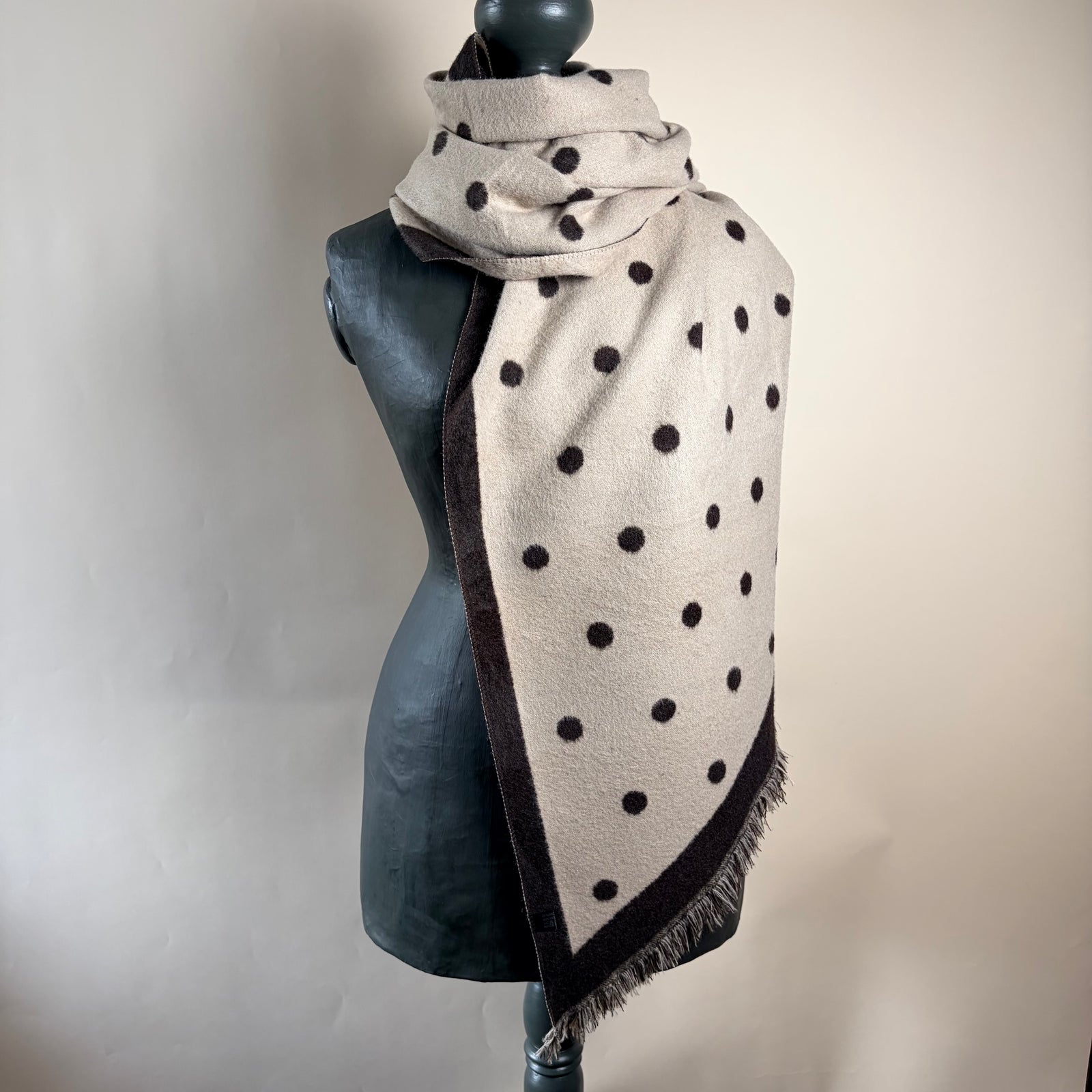 A dark brown and beige woolen scarf with white polka dots and fringed edges, draped on a black mannequin torso against a light beige background.