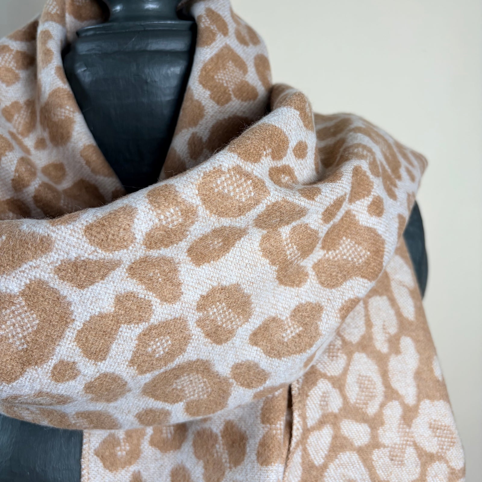A leopard print scarf in beige and brown, draped on a black mannequin torso against a light beige background.
