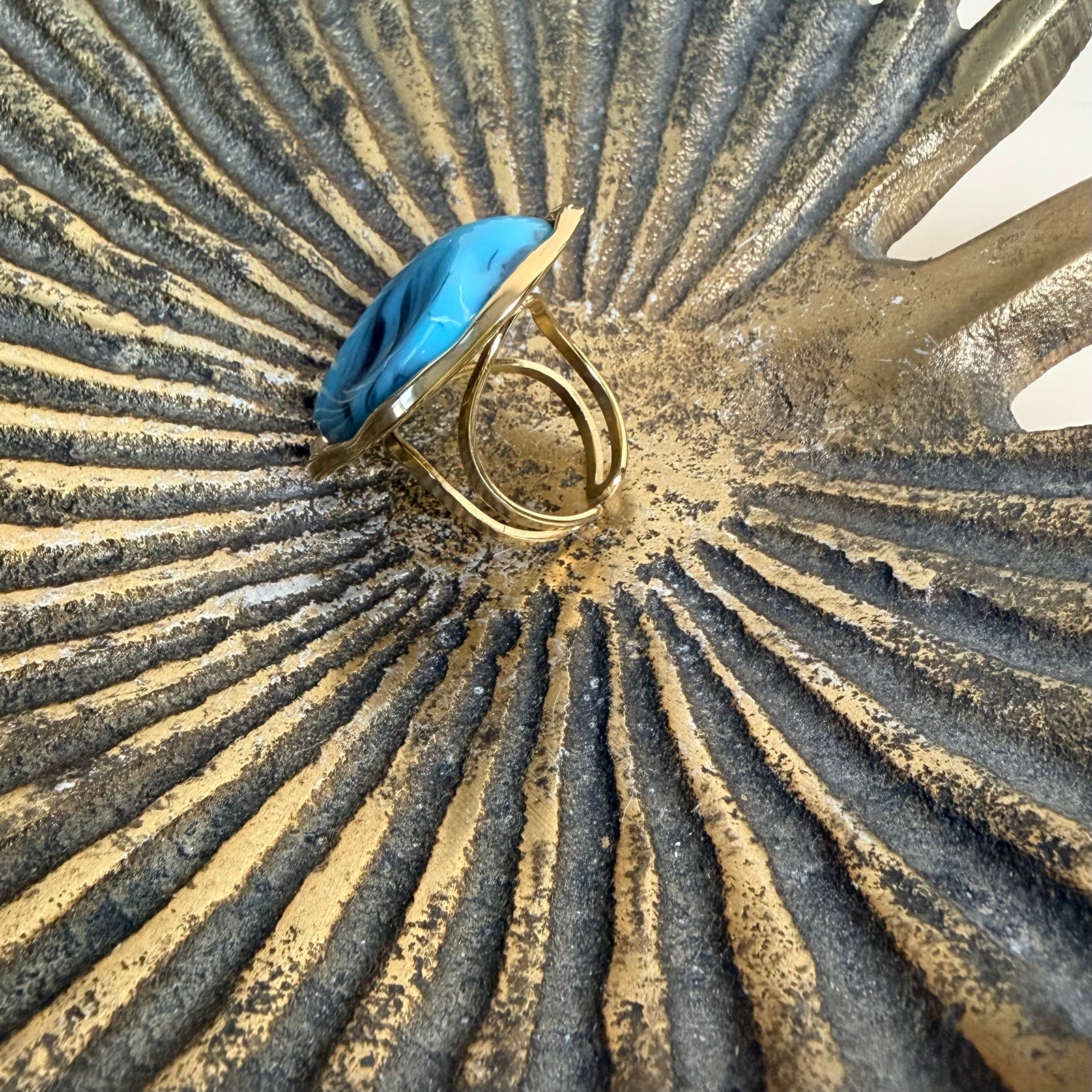 A ring featuring a large, irregular-shaped blue stone with dark marbled veins, set in a gold-toned bezel, displayed on a textured metallic gold and silver sunburst surface.
