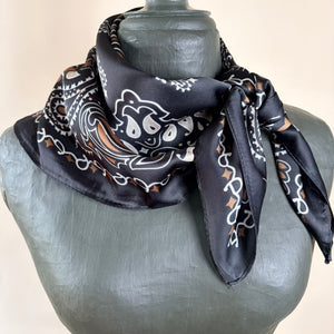 A black silk bandana scarf with a white and brown paisley pattern, styled on a gray mannequin bust against a light beige background.