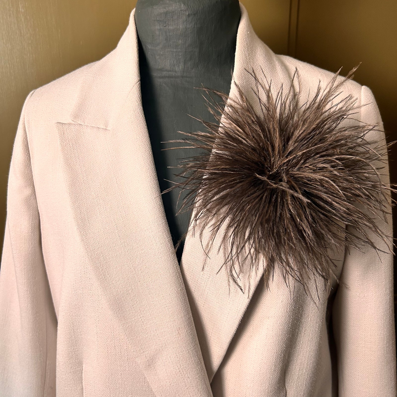 Mannequin wearing a beige blazer with a large brown feathered accessory on a plain background