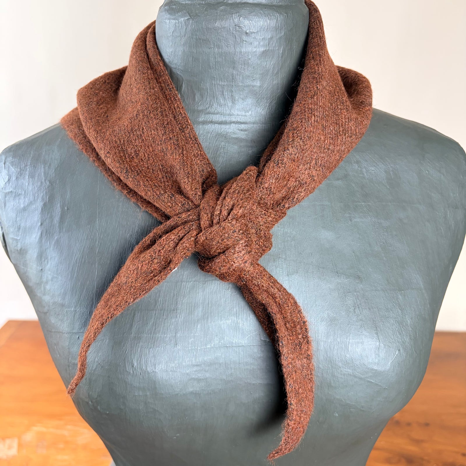 A brown textured knit scarf tied in a knot and draped around a gray mannequin torso, set against a neutral indoor background.