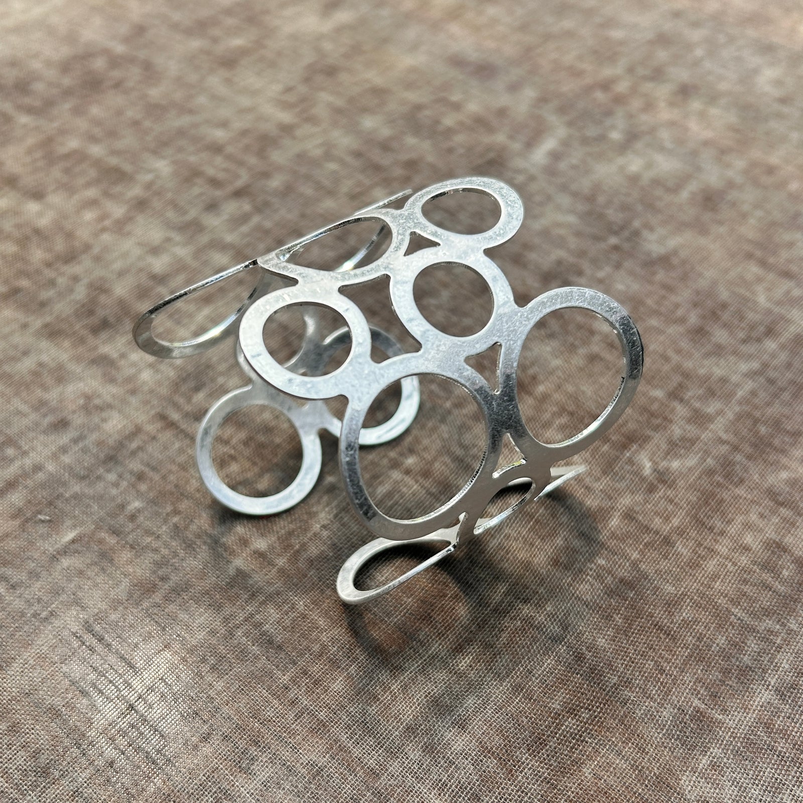 A metal geometric adjustable cuff bracelet with a silver finish, displayed on a textured background.