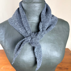 A gray knit scarf tied around a dark gray mannequin bust, with a soft neutral background and a wooden surface visible at the bottom.