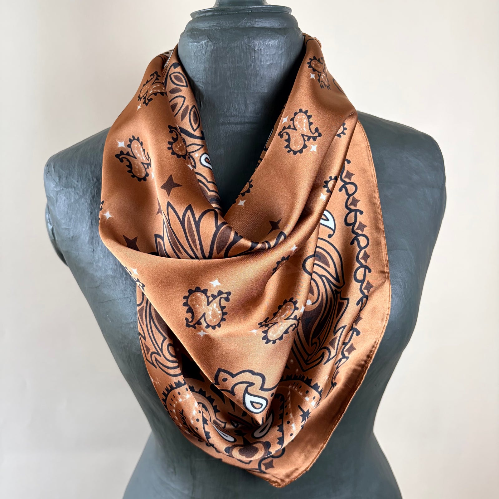 A brown satin scarf with a black and white paisley pattern draped on a dark gray mannequin torso against a light beige background.