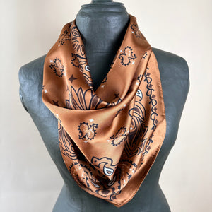 A brown satin scarf with a black and white paisley pattern draped on a dark gray mannequin torso against a light beige background.