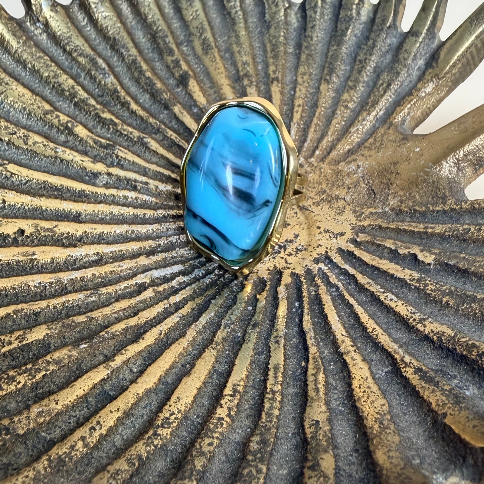 A ring featuring a large, irregular-shaped blue stone with dark marbled veins, set in a gold-toned bezel, displayed on a textured metallic gold and silver sunburst surface.