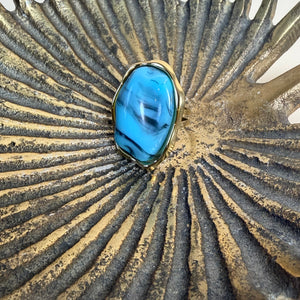 A ring featuring a large, irregular-shaped blue stone with dark marbled veins, set in a gold-toned bezel, displayed on a textured metallic gold and silver sunburst surface.